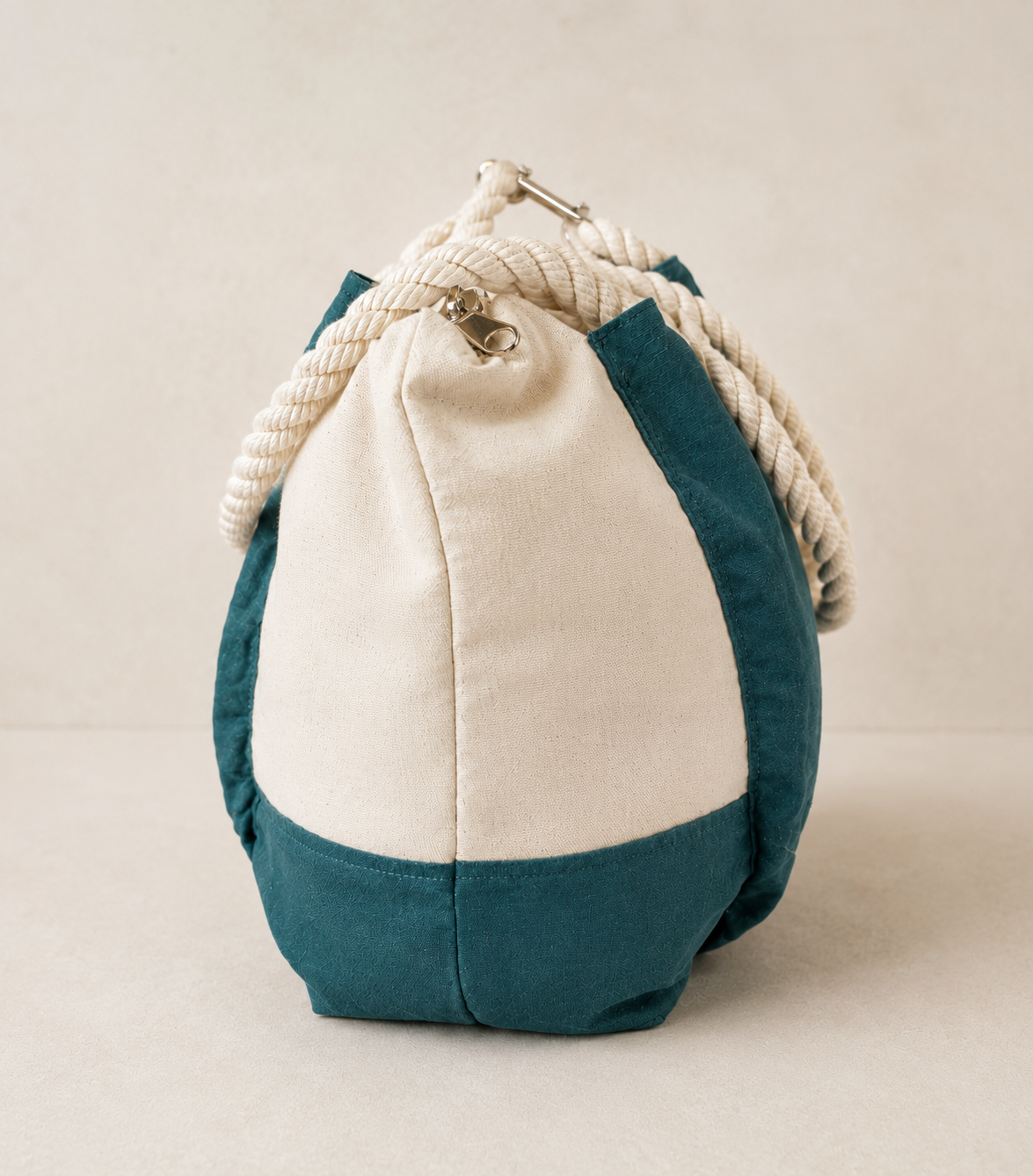 Side view of a natural canvas drawstring bag with teal contrast panels, thick rope handles, and silver hardware in a relaxed nautical-inspired silhouette.