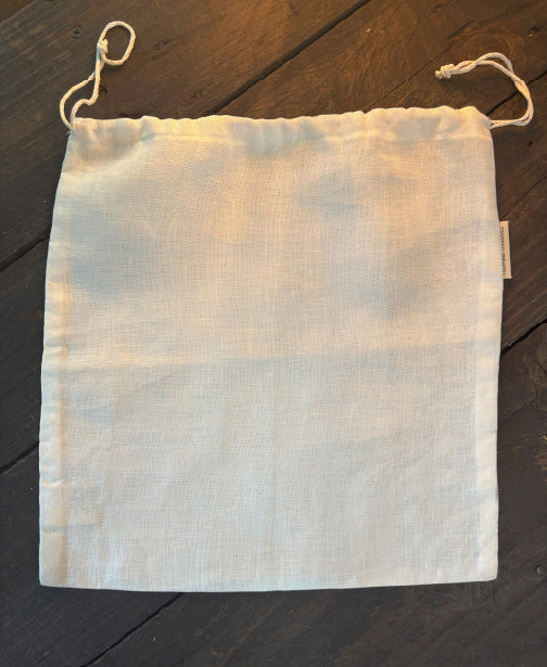 Natural flax linen bread bag with cotton drawstring, sized to hold a round or oblong loaf. Light, breathable fabric with a soft drape, photographed on a rustic kitchen surface with fresh bread partially visible inside.